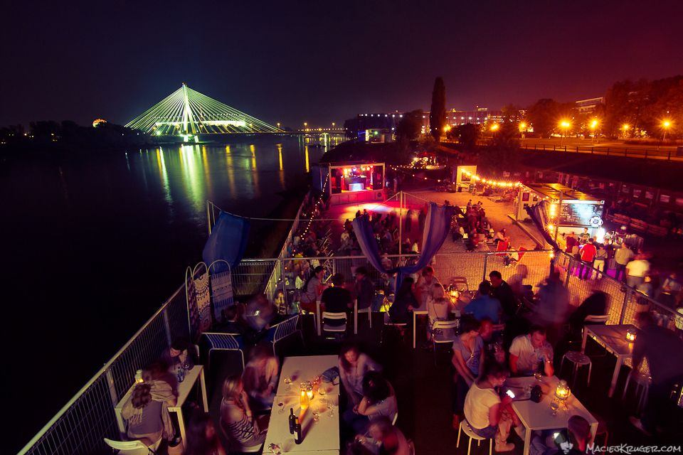 Outdoor riverside party venue in Warsaw at night with a lively crowd, city lights, and the Świętokrzyski Bridge in the background, Warsaw party calendar