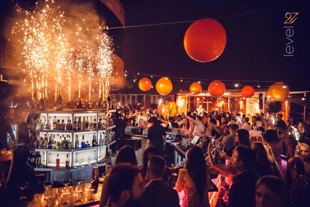 Crowded rooftop bar celebration in Warsaw with champagne bottles, fireworks, and glowing lanterns during a premium night event, Warsaw party calendar