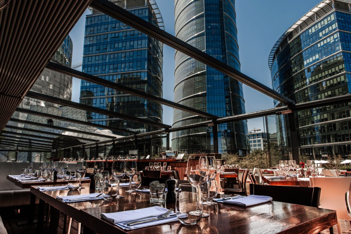 Elegantly set wooden dining tables with wine glasses on an outdoor restaurant terrace under a glass roof, surrounded by towering modern glass skyscrapers on a sunny day.