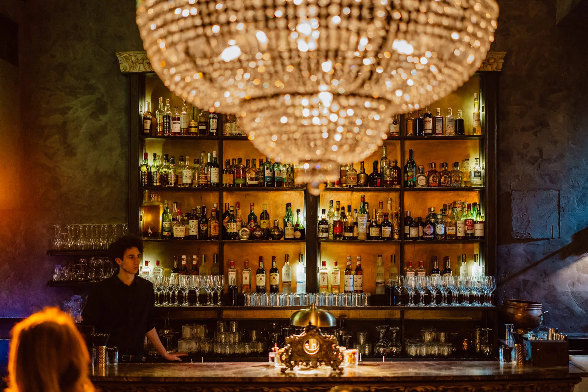 The dimly lit interior of Weles Bar featuring a massive, glowing crystal chandelier hanging over a wooden bar counter fully stocked with liquor bottles.